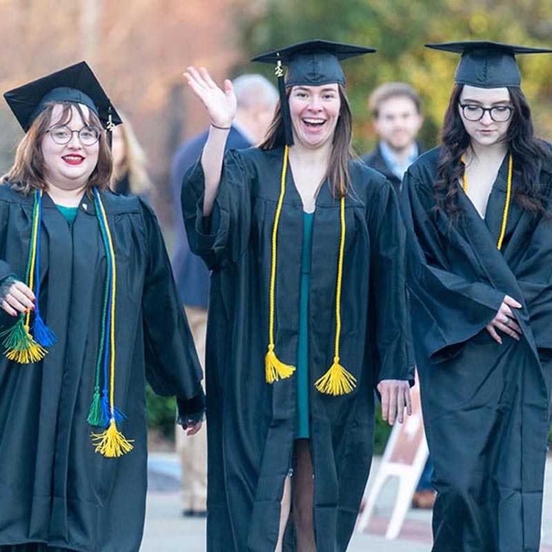 Students in cap &  gowns smile and wave to camera as they walk to their graduation ceremony in Curb Arena 