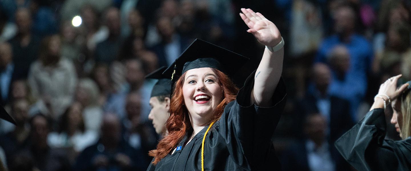 Graduate waving to family member in crowd while walking into graduation ceremony