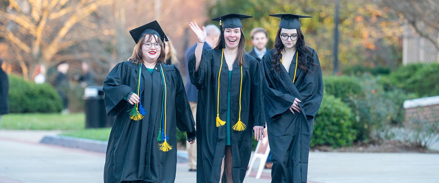 Students in cap &  gowns smile and wave to camera as they walk to their graduation ceremony in Curb Arena 