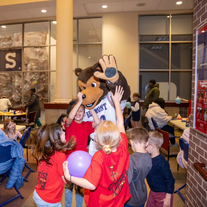Children joyfully gather around Bruiser, Belmont mascot, wearing a "Belmont" jersey. Lively and playful atmosphere with others seated nearby.