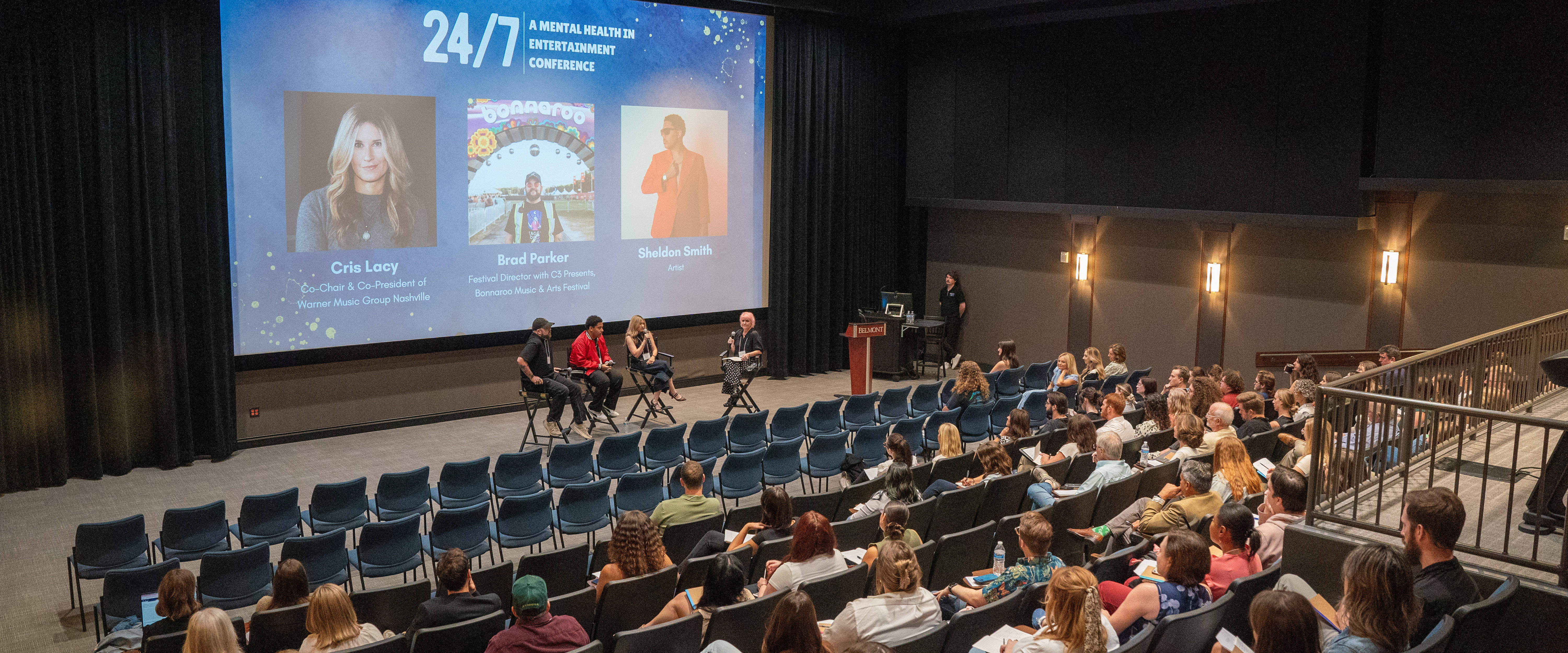 Panelists speak onstage during the “24/7: A Mental Health in Entertainment Conference” at Belmont University. The large screen behind them displays headshots and names of featured guests: Cris Lacy (Co-Chair & Co-President, Warner Music Group Nashville), Brad Parker (Festival Director, Bonnaroo Music & Arts Festival), and Sheldon Smith (Artist). The audience sits attentively in a modern auditorium with stadium-style seating.