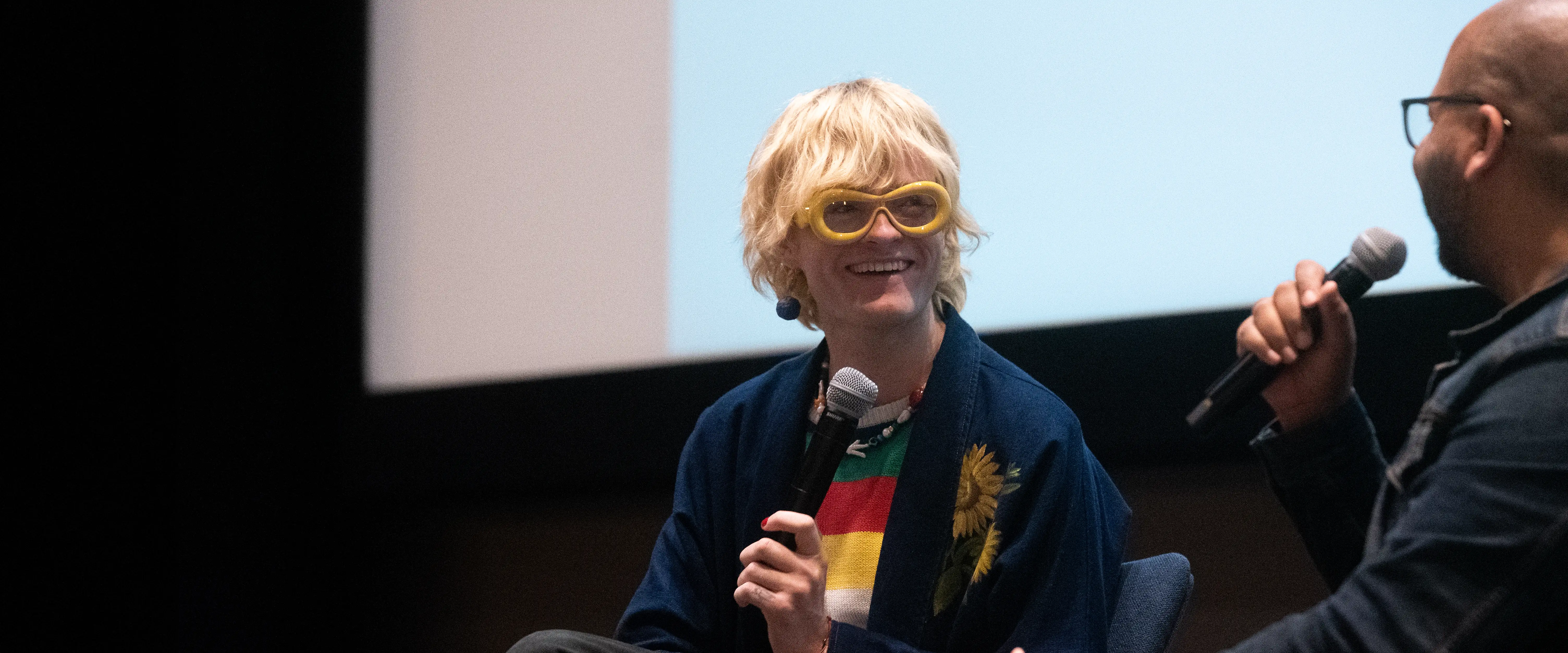 Belmont Alum Jake Wesley Rogers and interviewer engaged in a panel discussion on stage. The person on the left, wearing yellow oversized glasses, a blue jacket with sunflower embroidery, and a colorful striped shirt, holds a microphone and smiles. The person on the right, partially visible, wears glasses and a dark jacket while speaking into a microphone. A large projection screen is visible in the background.