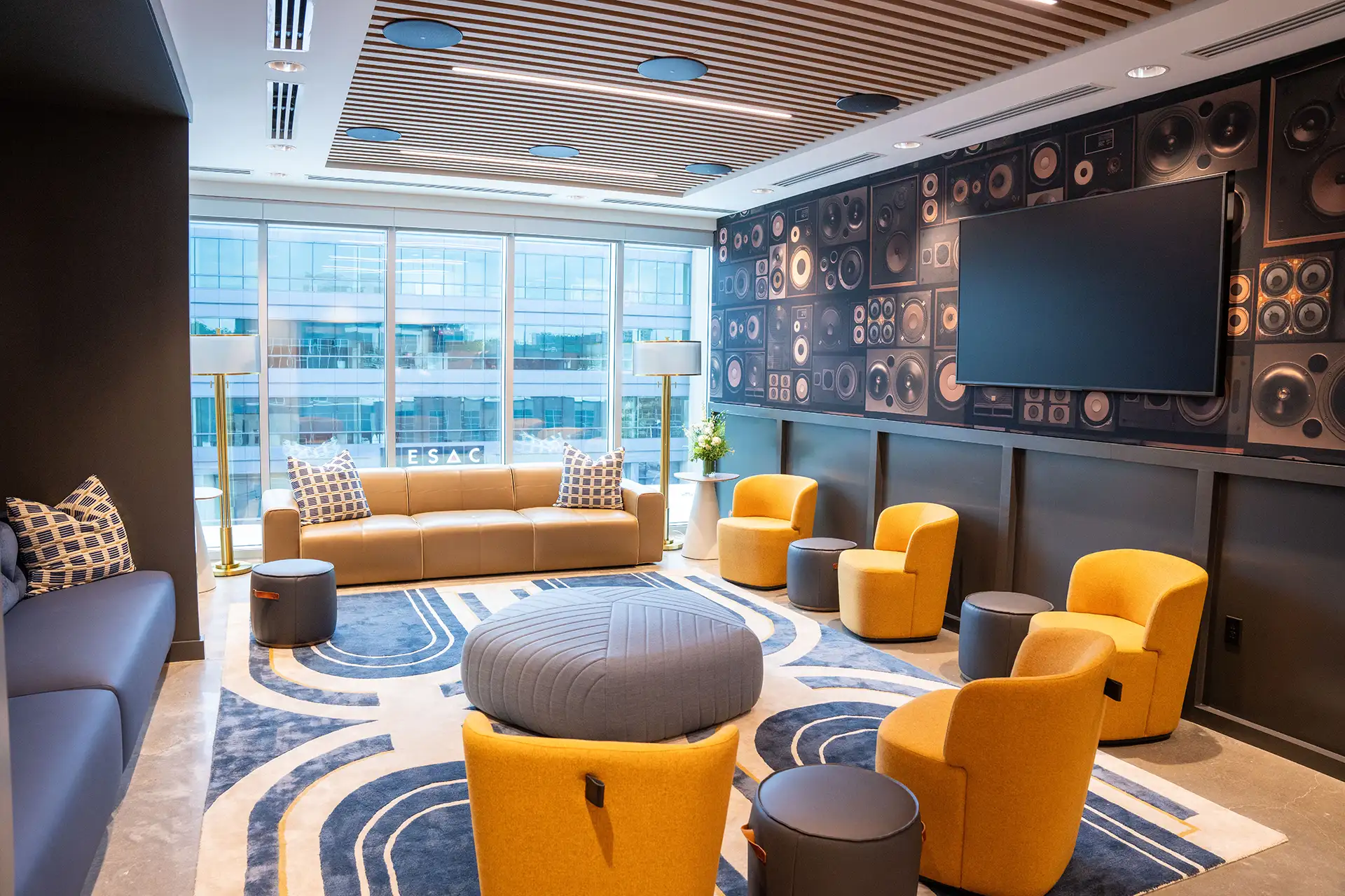 Bright lounge with yellow and gray chairs, large windows, patterned rug, and a wall decorated with speaker graphics.