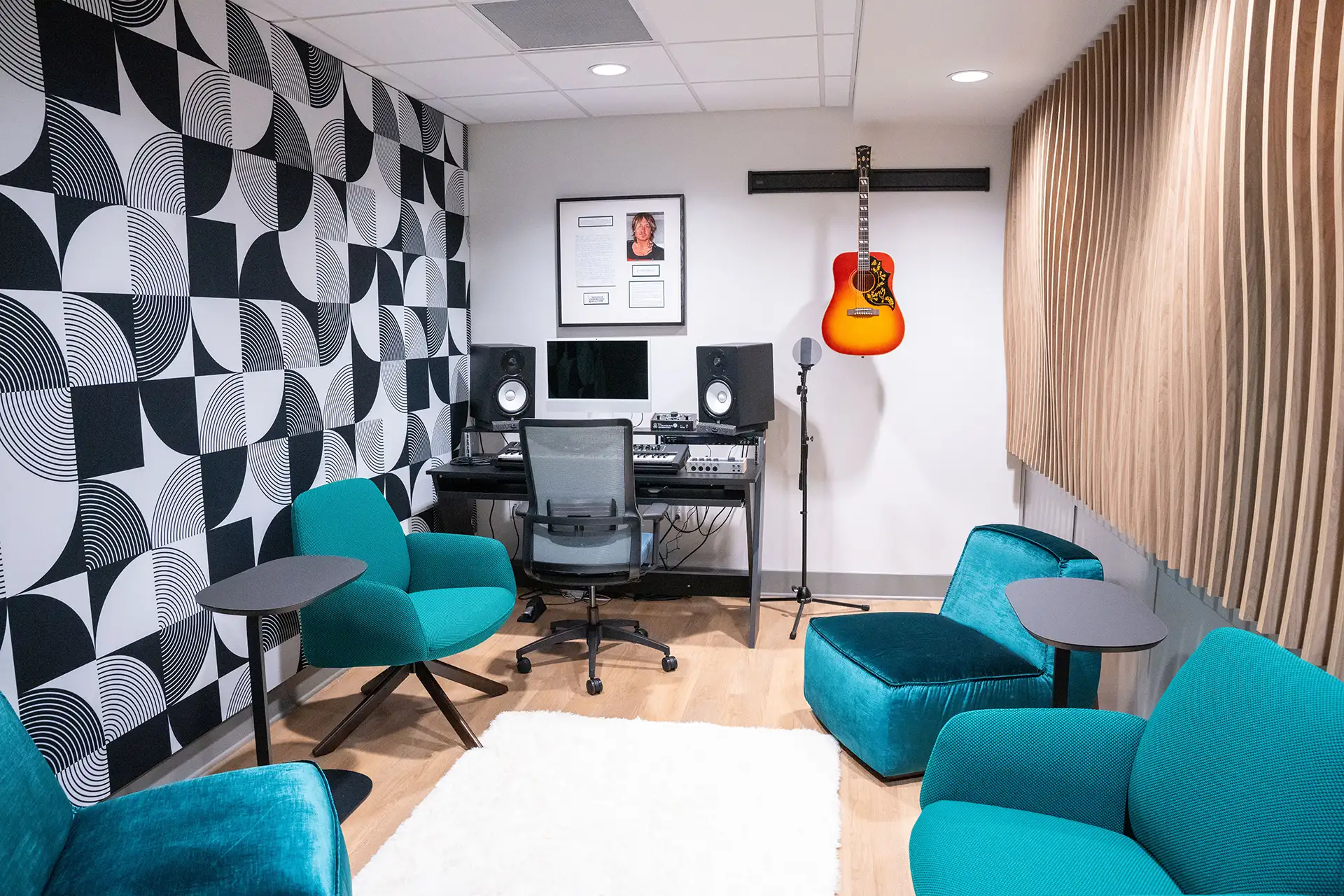Small studio room with teal lounge chairs, patterned black-and-white wall, sound equipment, and a mounted guitar.