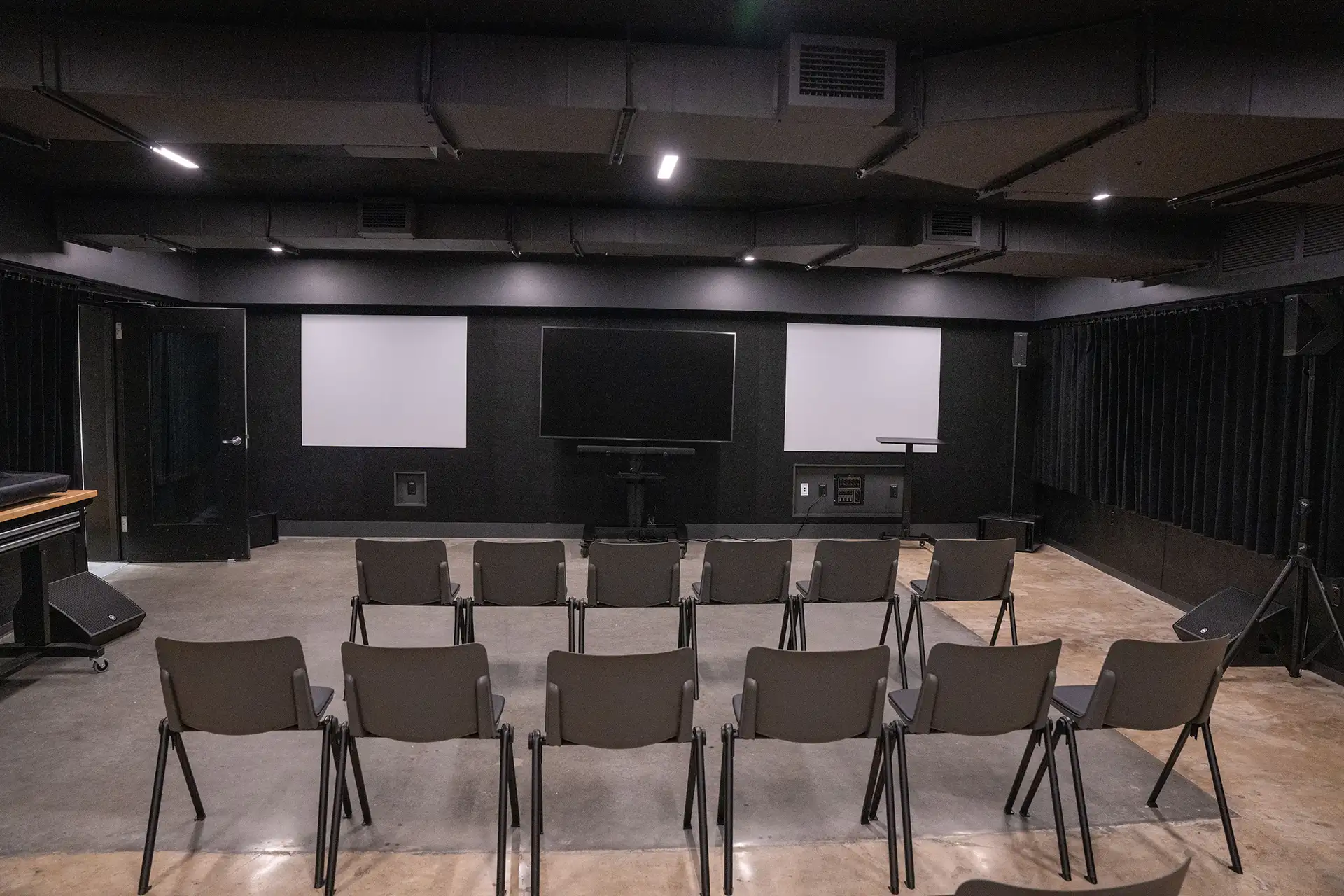 Black box theater setup with rows of chairs facing multiple projection screens and a central TV.