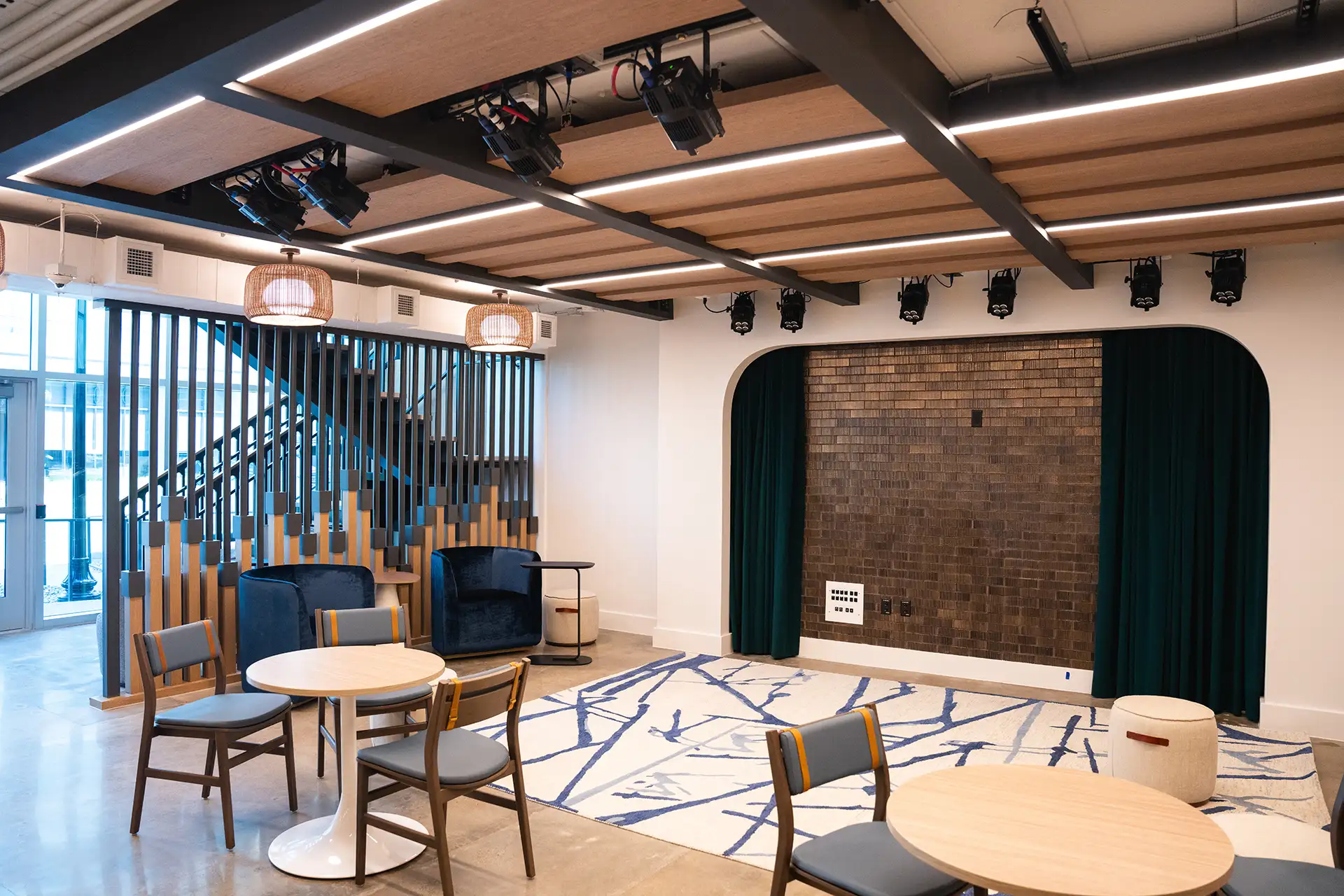 Small performance space with round tables, blue chairs, stage lighting, and a brick backdrop.