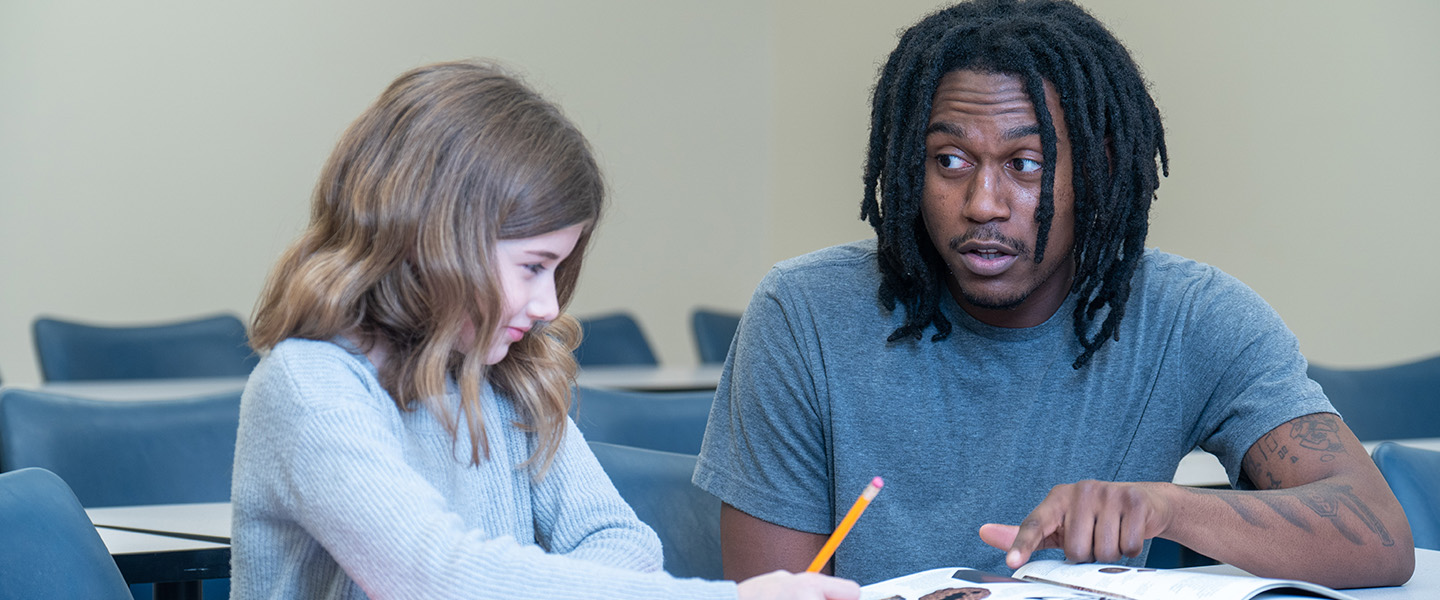 A male teacher sits with a young student at a desk and directs her attention to a book.