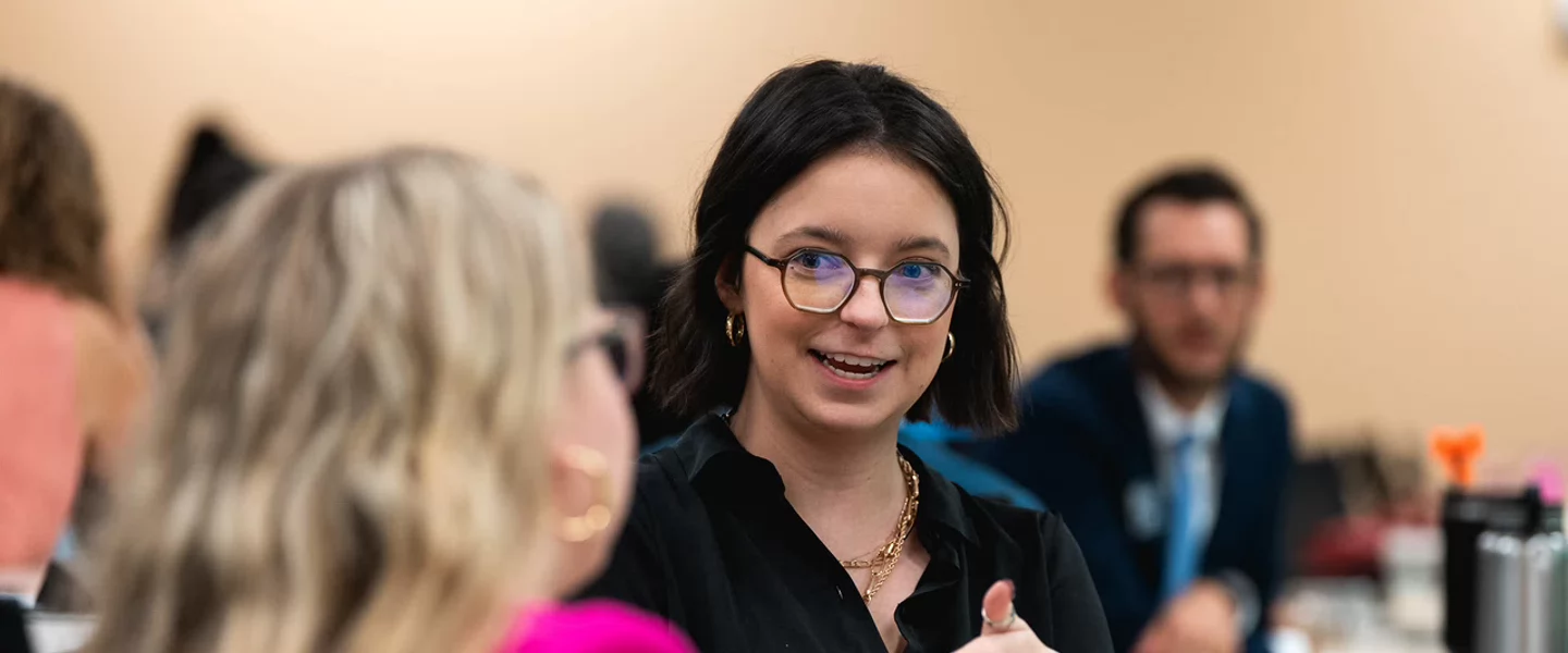 A woman wearing glasses and black professional attire smiles while speaking at a meeting or classroom setting with other participants visible in the background.