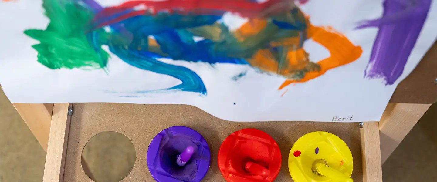 A close-up view of a child's artwork displayed on a wooden easel, featuring vibrant brush strokes in various colors. Below the artwork, three paint containers in purple, red, and yellow are visible, each with a paintbrush inserted, indicating an engaging creative activity.