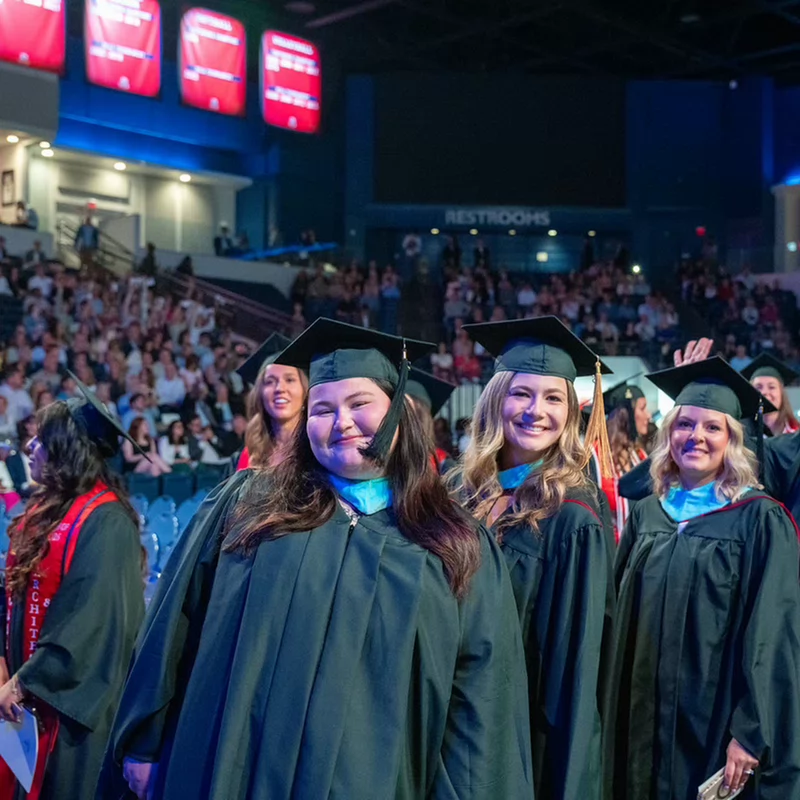 Belmont University graduates in black caps and gowns stand together smiling during their graduation ceremony in an arena filled with the audience.