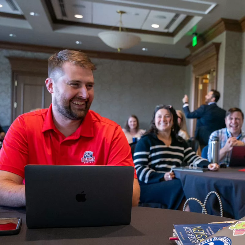 A man wearing a red Belmont polo shirt smiles while working on a laptop at an event, with a woman beside him and other participants visible in the background.