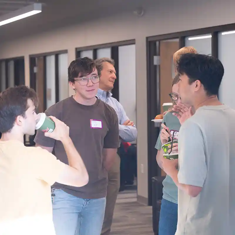 A group of students and faculty socialize in a hallway, enjoying refreshments and conversation.