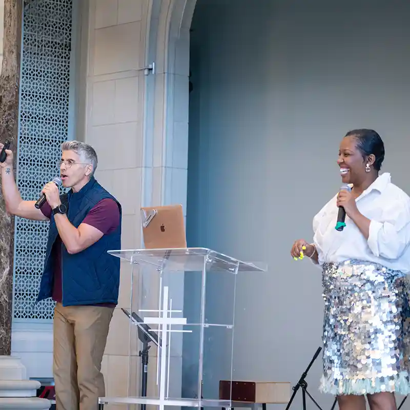 Two speakers hold microphones during a chapel gathering, standing beside a clear podium with a cross.