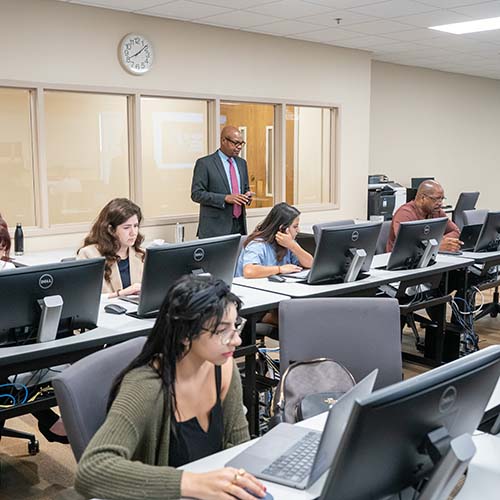 Students working on classwork at computers while professor lectures the class