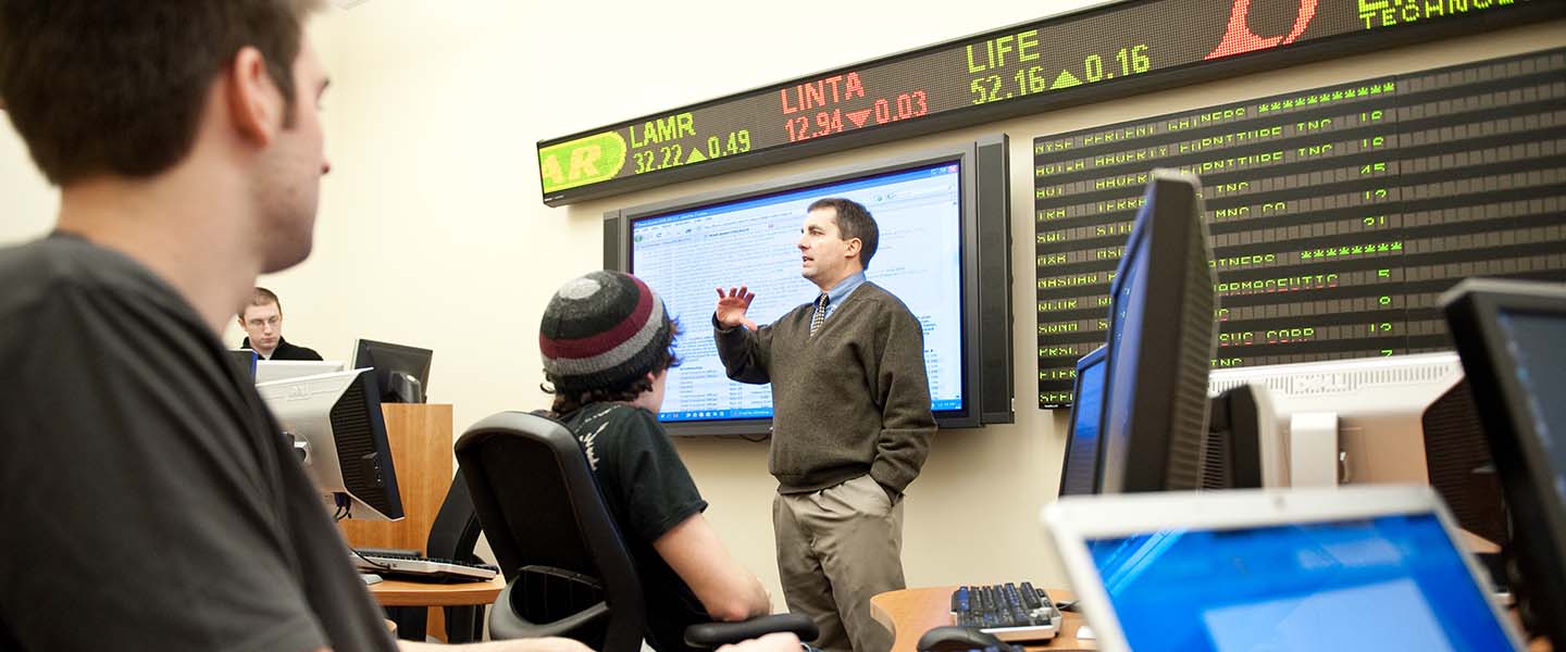 Professor teaches in front of class while students take notes on their laptops