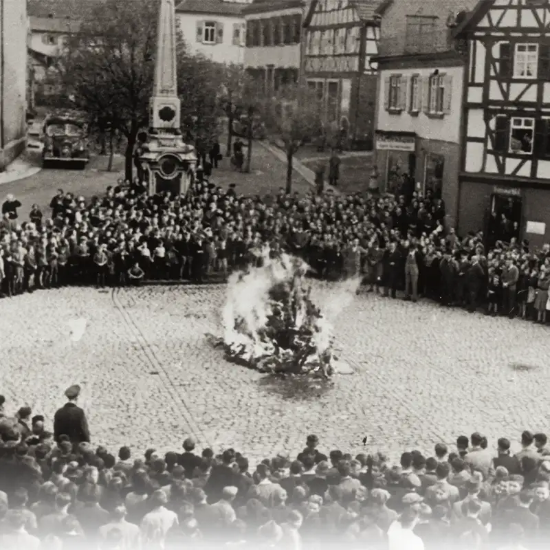 A historical black-and-white photograph showing a large crowd gathered around a bonfire in a cobblestone square. Buildings in the background are visible, along with trees. The scene depicts people observing the fire, some standing while others are seated.