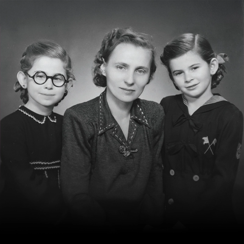 Black and white portrait of a woman seated between two young girls; the girl on the left wears round glasses and a dark dress, while the girl on the right wears a sailor-style outfit.