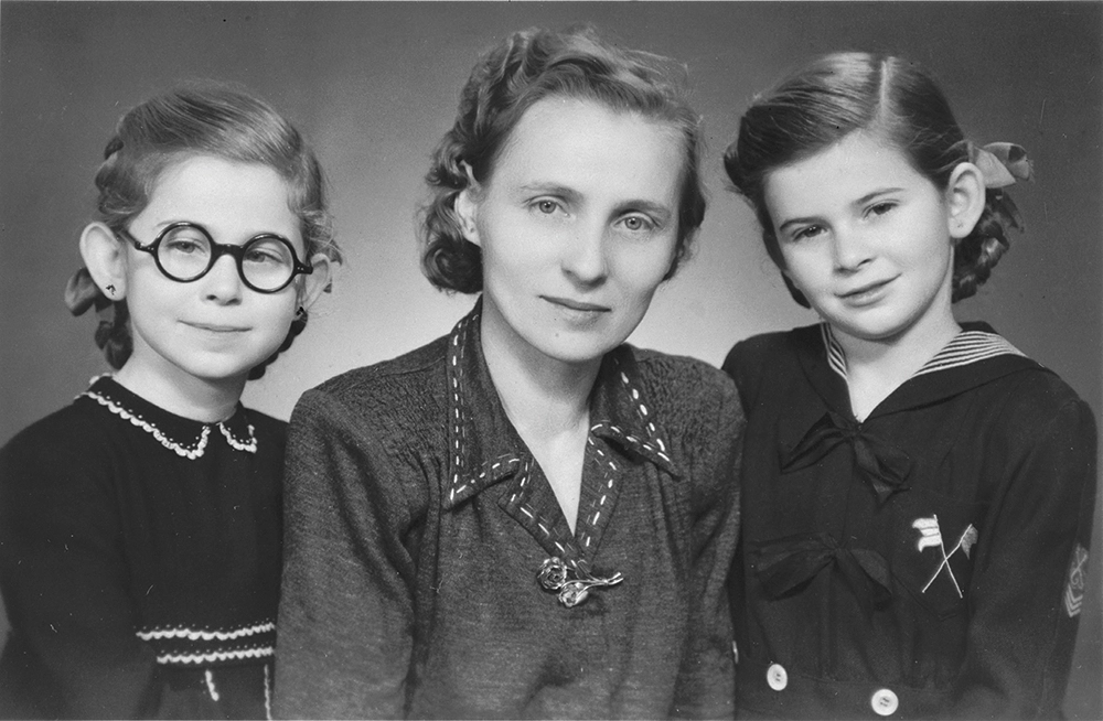 A black-and-white portrait of a woman and two young girls. The woman is seated in the center, with the girls standing on either side of her. One girl wears round glasses and a dark dress with light trim, while the other wears a sailor-style outfit. All three look calmly toward the camera.