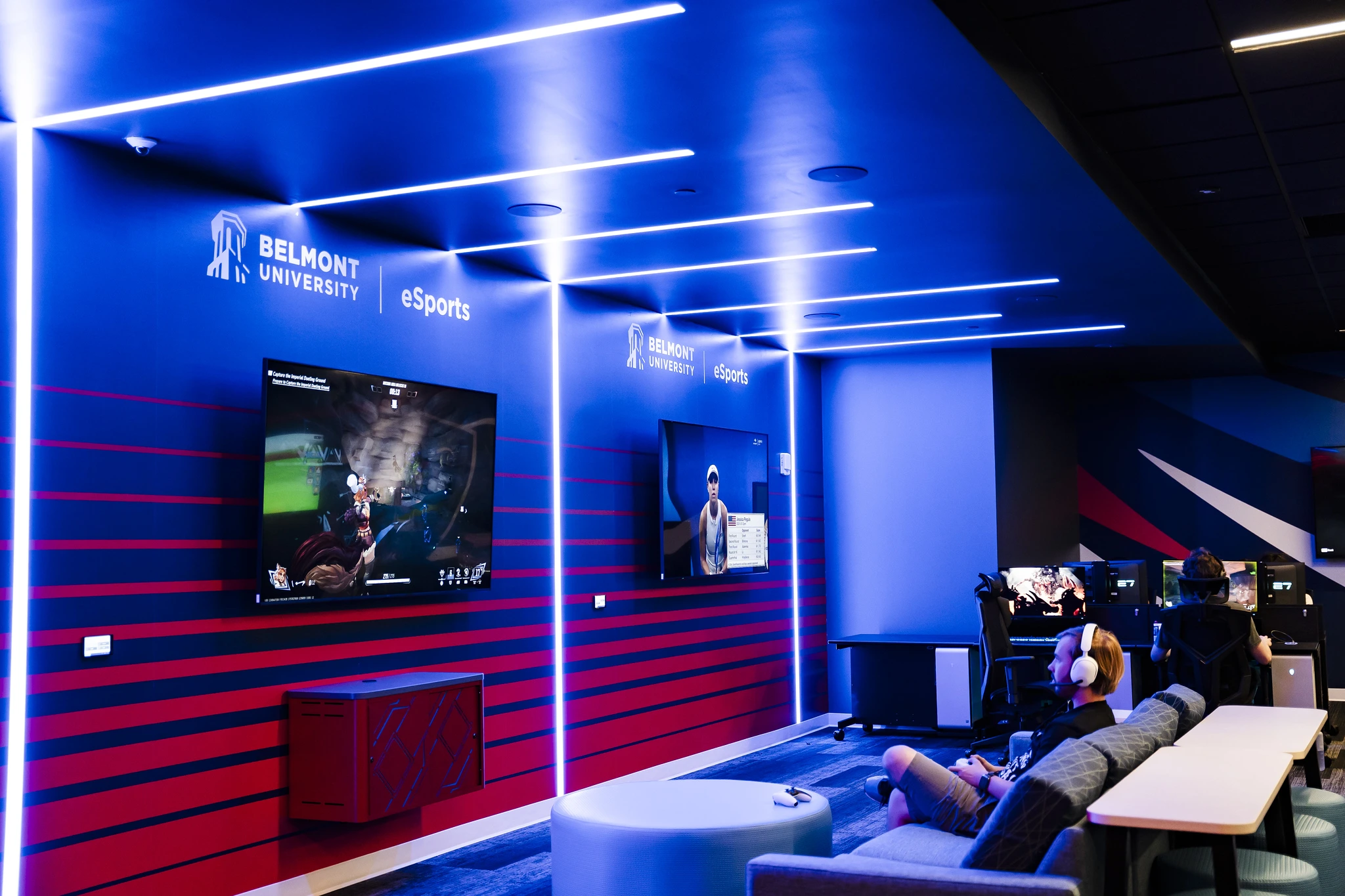Belmont University eSports gaming lounge with blue LED ceiling lights, wall-mounted game screens, and students playing at desktop PCs.