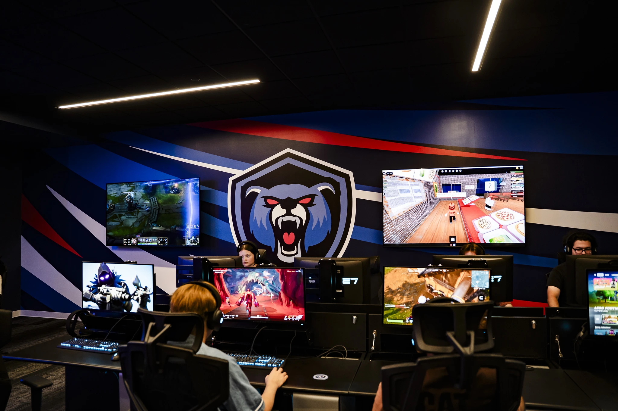 Gamers play on desktop PCs in an esports lounge, with large wall screens showing gameplay beneath a stylized bear team logo mural.