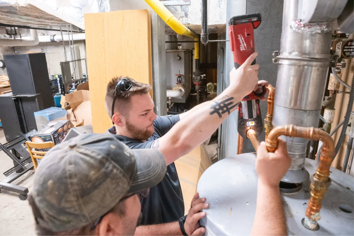 Two plumbers using a pipe press tool to install copper plumbing on a large water heater.