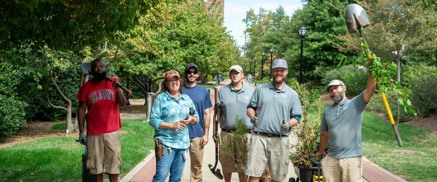 Six people stand on a tree-lined path, smiling and holding tools. A man raises a shovel triumphantly.