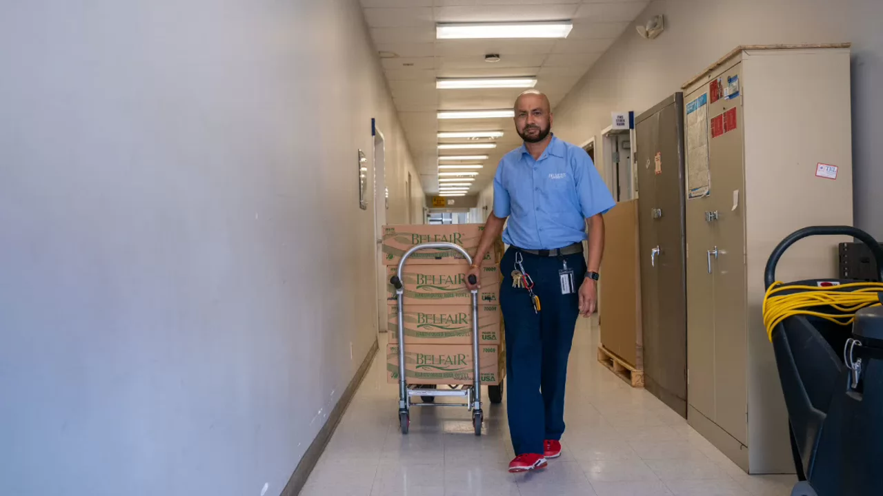 Belmont Custodial staff pushes a cart stacked with brown boxes down a long hallway, delivering supplies.