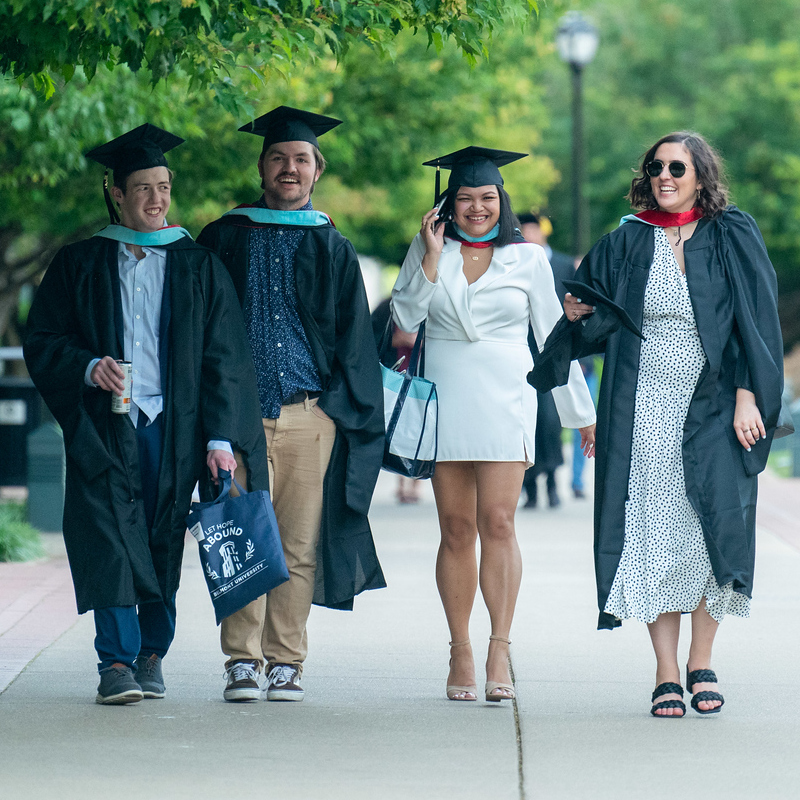 A group of four students in graduation regalia walking across campus towards commencement ceremony