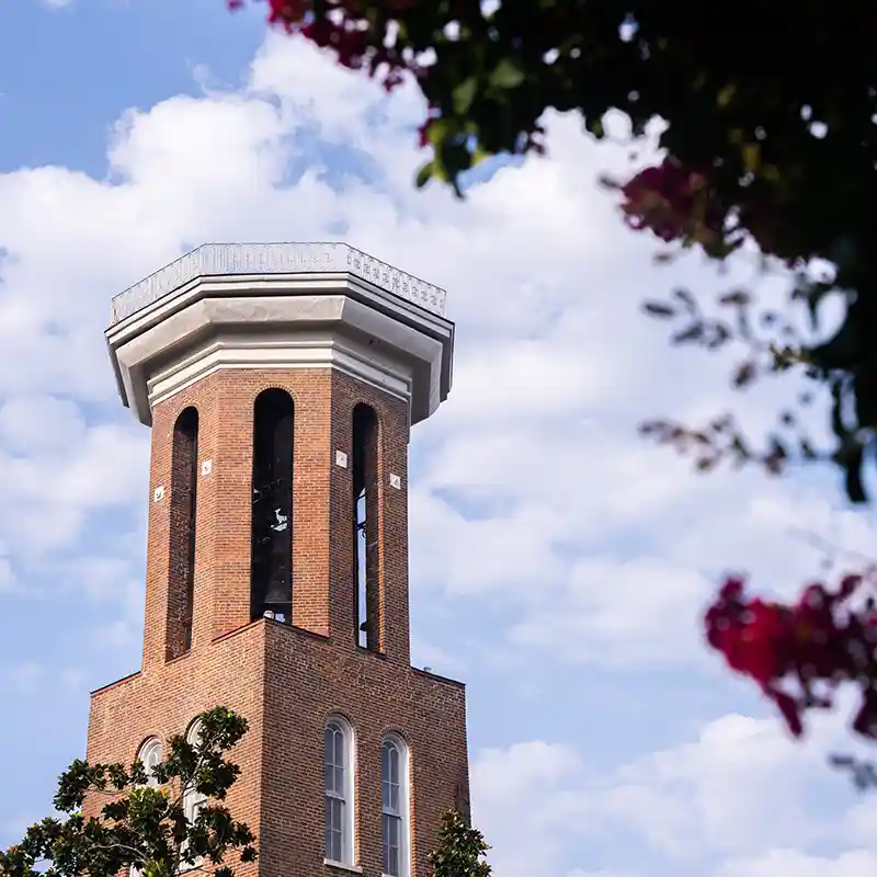 Belmont University's historic brick Bell Tower against a partly cloudy blue sky, framed by flowering trees.