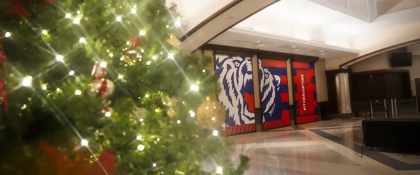 Christmas tree decorated with glowing lights and ornaments inside a Belmont University building, with a large Bruin graphic in the background.