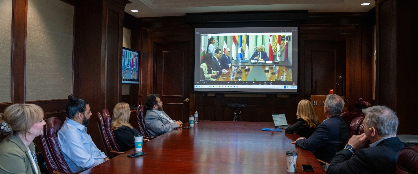 Executives in a board room watching a virtual international meeting displayed on a large projector screen.