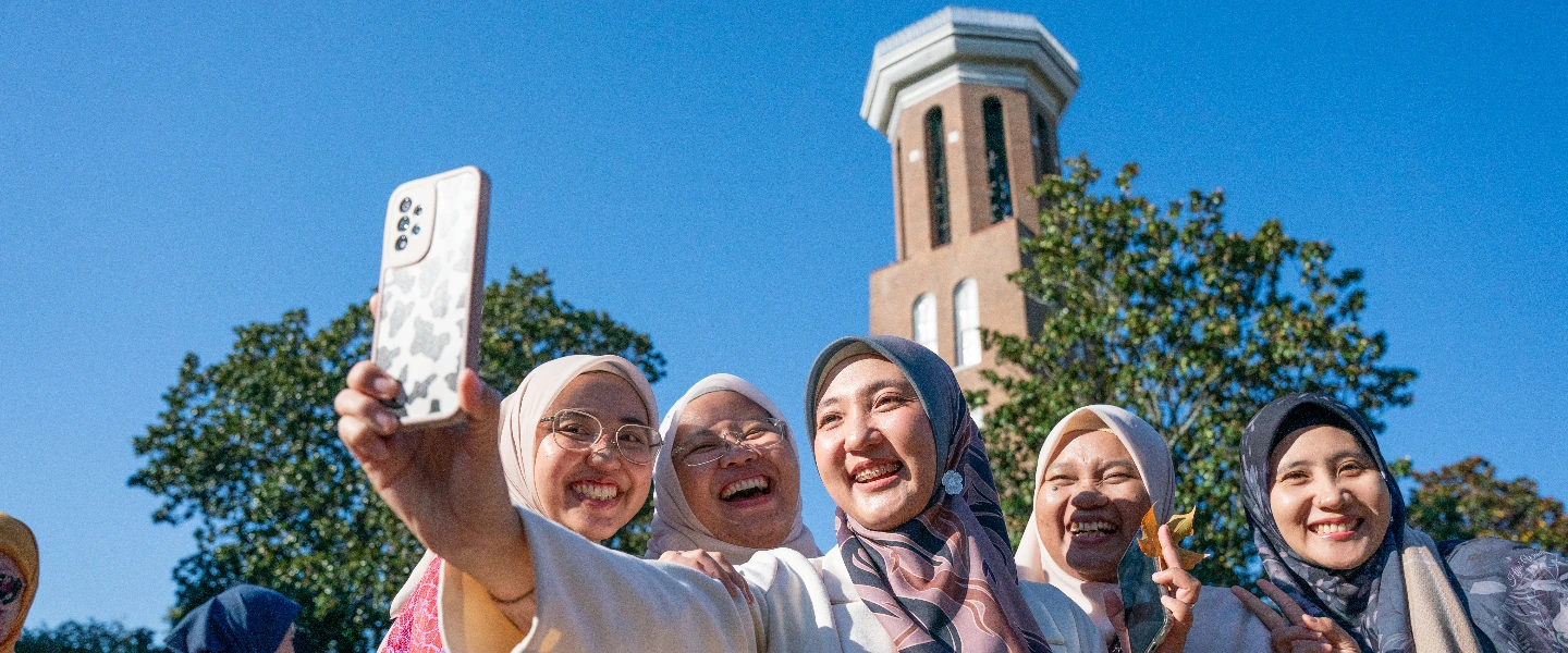 Five happy women in hijabs taking a selfie outdoors, with a tall brick tower and blue sky in the background.