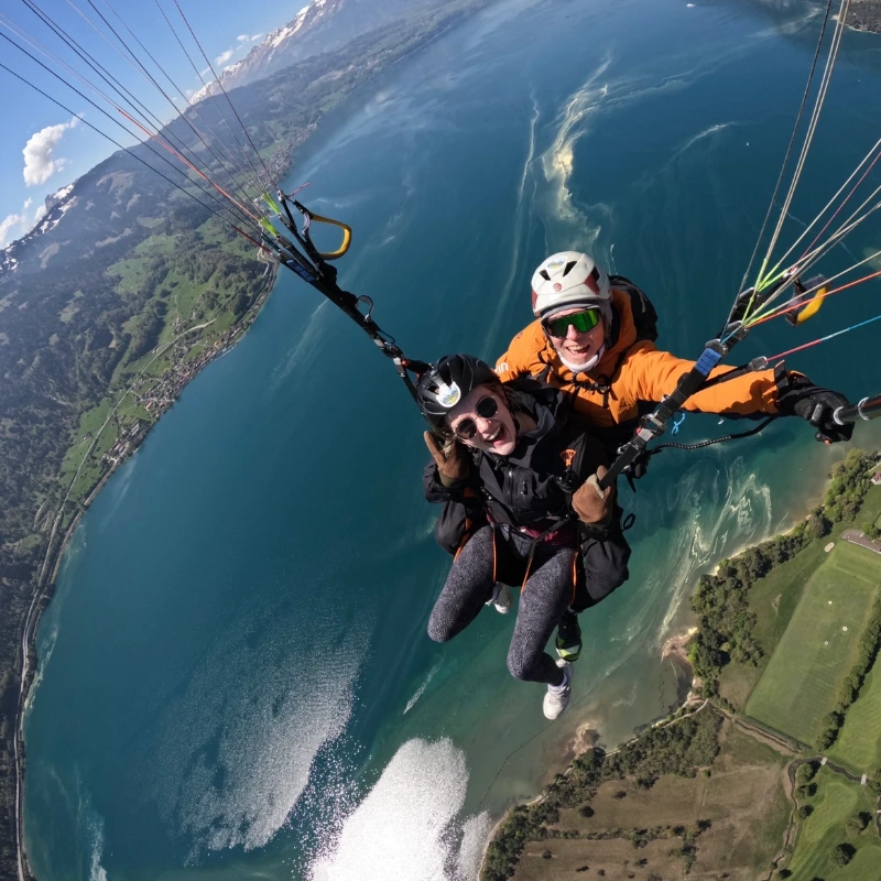 Smiling duo paragliding above a scenic blue lake and lush green mountains, thrilling adventure.