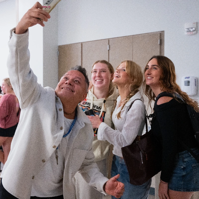 Happy man taking a selfie with three smiling young women, likely friends or students at an event.
