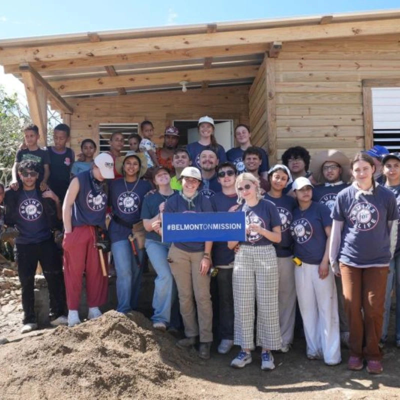 Belmont mission trip volunteers stand by a newly built wooden house, holding a #BELMONTONMISSION banner.