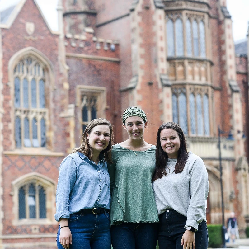 Three smiling young women stand outside a historic brick university building with gothic architecture.