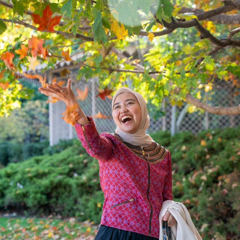 Happy woman in hijab throwing autumn leaves into the air, enjoying fall outdoors.