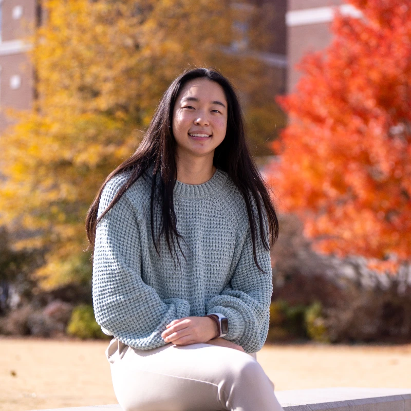 Young Asian woman smiling, wearing a light blue sweater, with vibrant autumn trees and a campus building in the background.