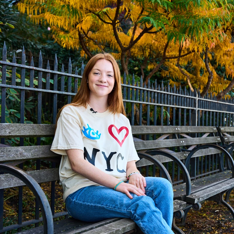 Young woman with red hair in an "I Love NYC" shirt smiles on a park bench with autumn trees.