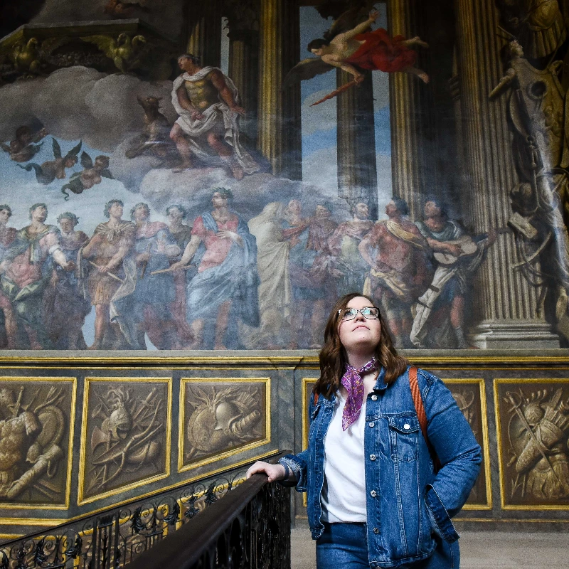 Woman in denim jacket on a grand staircase admiring a large, ornate classical fresco mural.