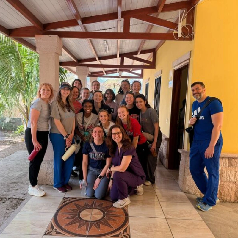Large group of smiling Belmont students and volunteers on a medical mission trip posing on a covered porch.