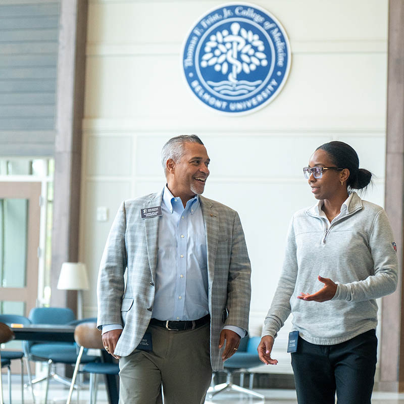 Two professionally dressed individuals walk and converse inside the Thomas F. Frist, Jr. College of Medicine at Belmont University. A large Frist College of Medicine seal is visible on the wall behind them.