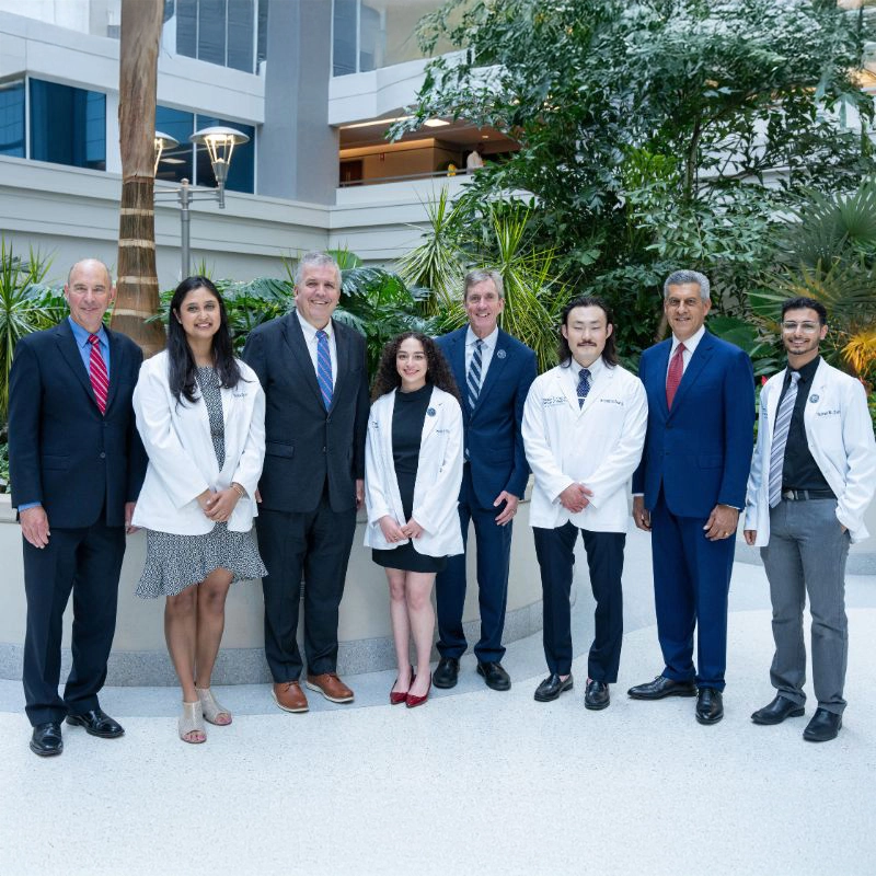 A group of eight people, including four individuals in white medical coats and four in business attire, stand together indoors in a bright, modern space with tropical plants in the background.
