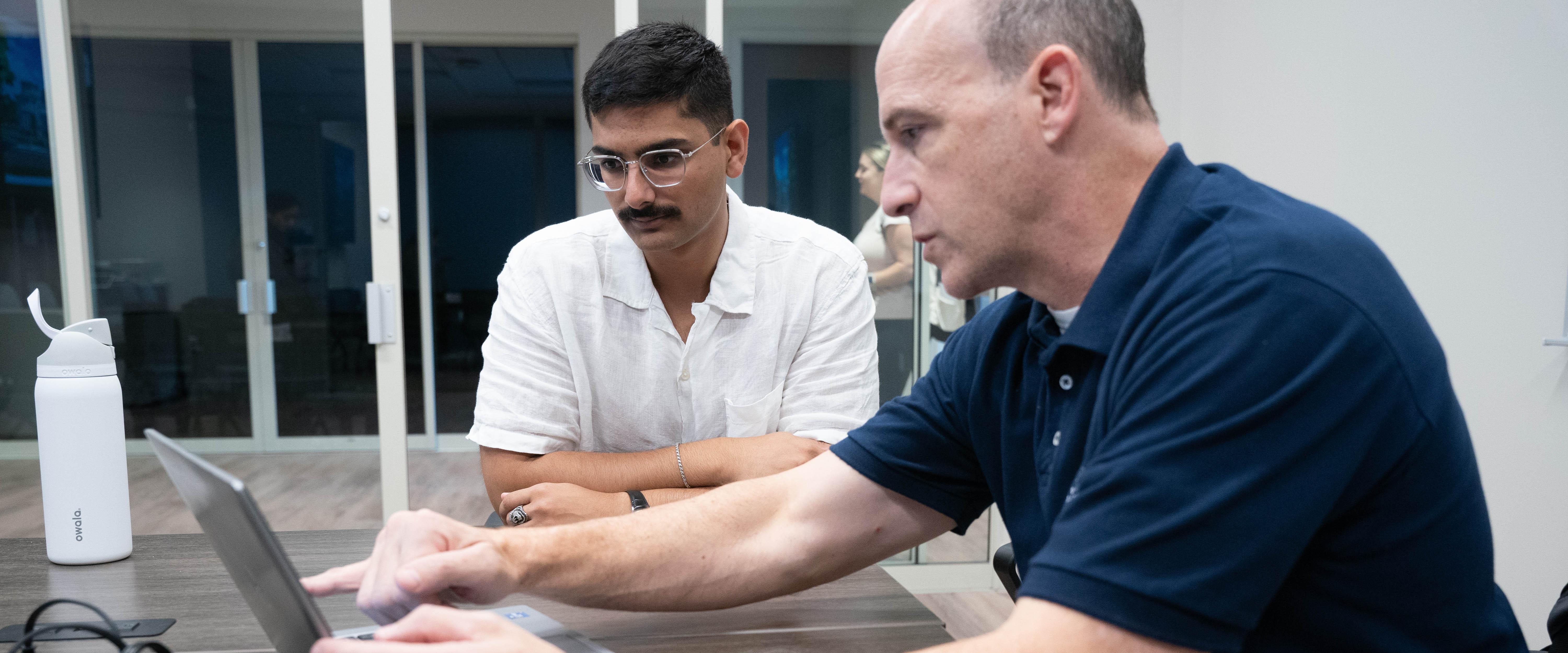 A man in a navy blue polo shirt is pointing at a laptop screen while explaining something to another man in a white shirt with glasses and a mustache. They are seated at a conference table in a modern office setting with glass doors in the background.