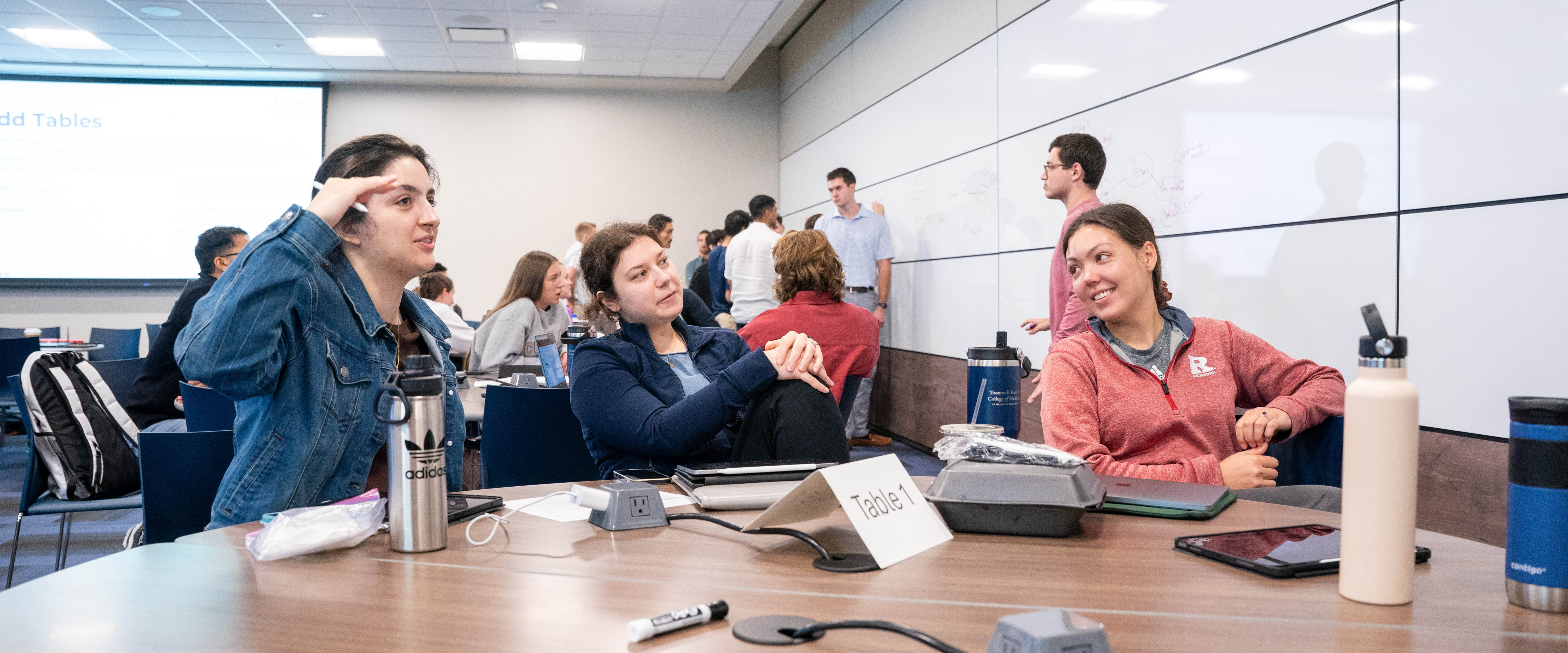 A group of young professionals is engaged in a discussion in a classroom or workshop setting. Three women at the front table are conversing and smiling, while other participants work and write on whiteboards in the background