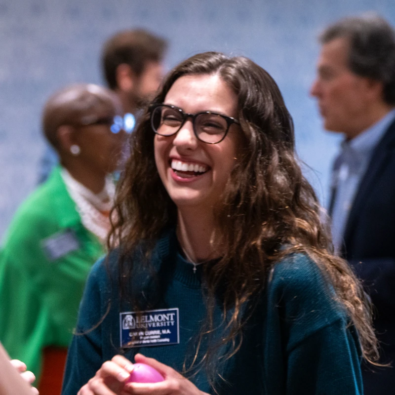 A woman with long brown hair and glasses smiles while holding a pink object at an indoor event with other people in the background.