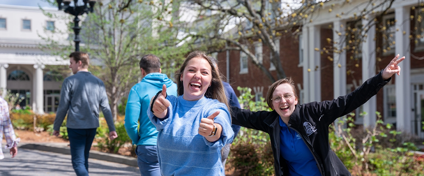 Two joyful women walking on Belmont University campus, one giving thumbs-up, the other with arms wide, smiling broadly.