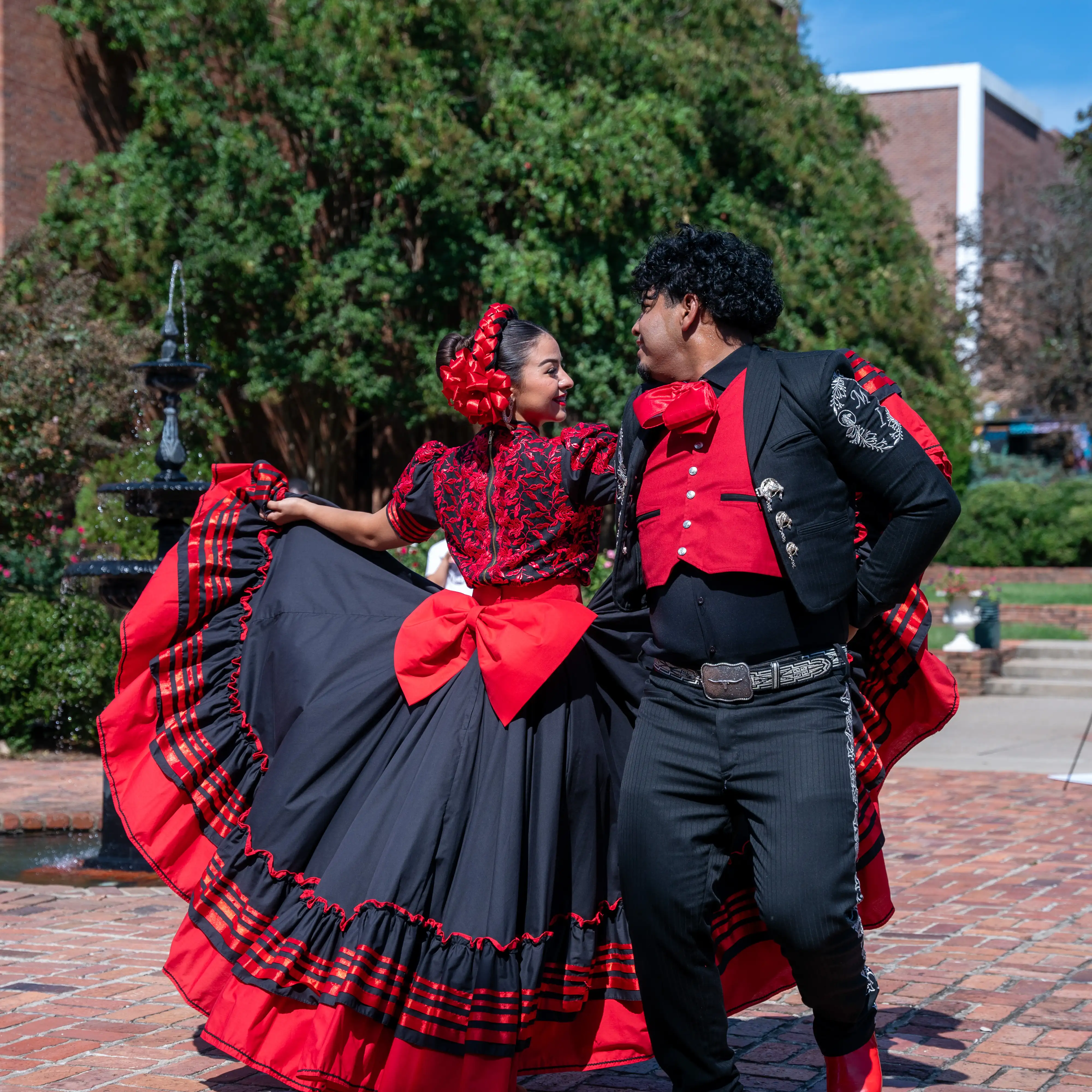 Two student dancers perform a traditional folklórico-style dance in red and black costumes on Belmont’s campus, posing mid-movement near a brick plaza and fountain.