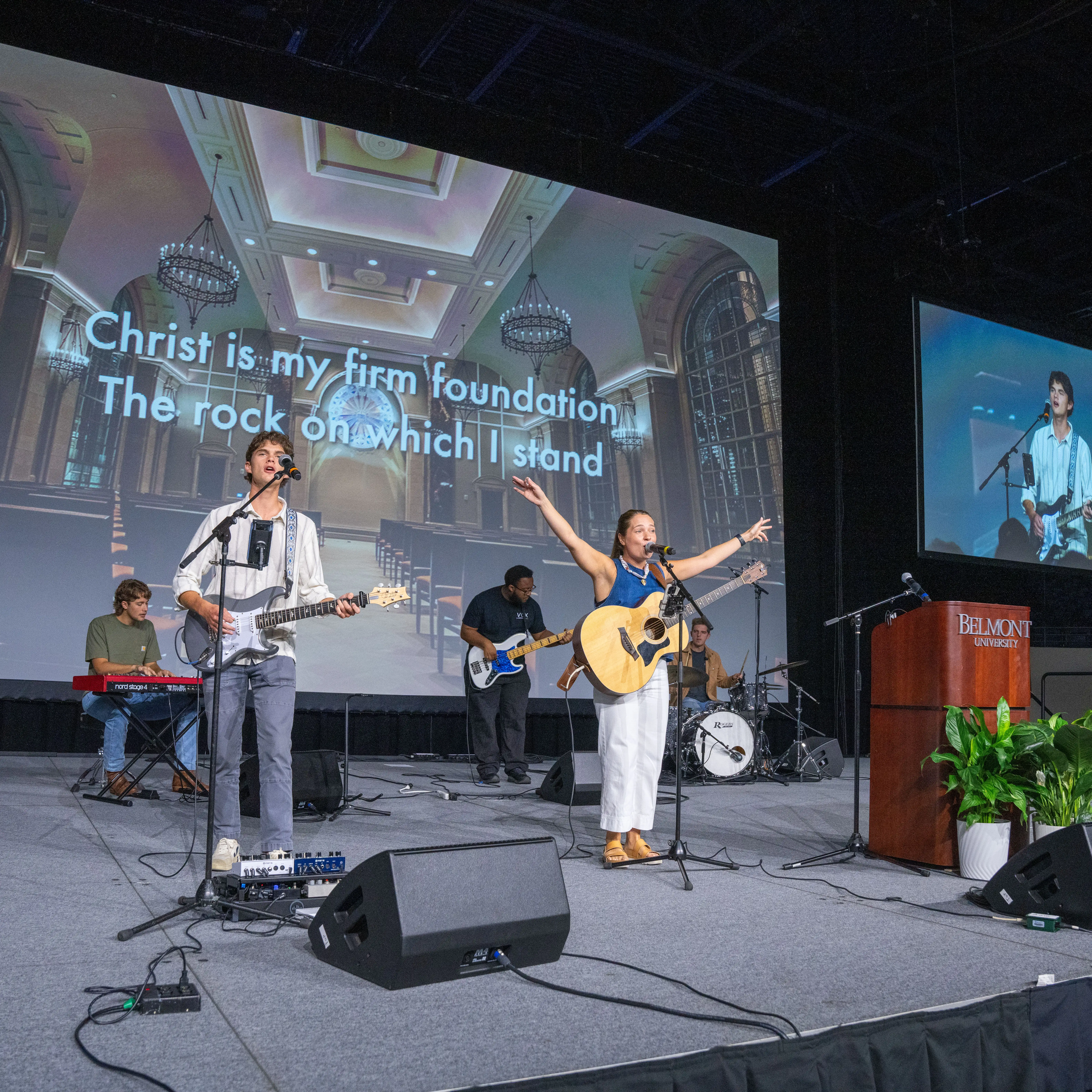 Students lead a worship service on a stage at Belmont University, singing and playing instruments beneath projected lyrics inside a large indoor venue.
