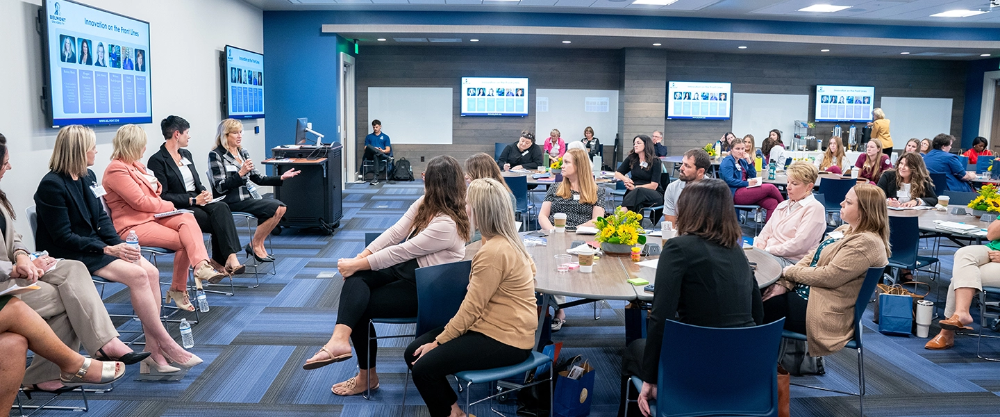  A panel of women speaks to a seated audience in a modern conference room during a professional event at Belmont University. The screens display the session title, “Innovation on the Front Lines,” and headshots of panelists. Attendees sit at round tables with coffee and notepads, listening attentively.